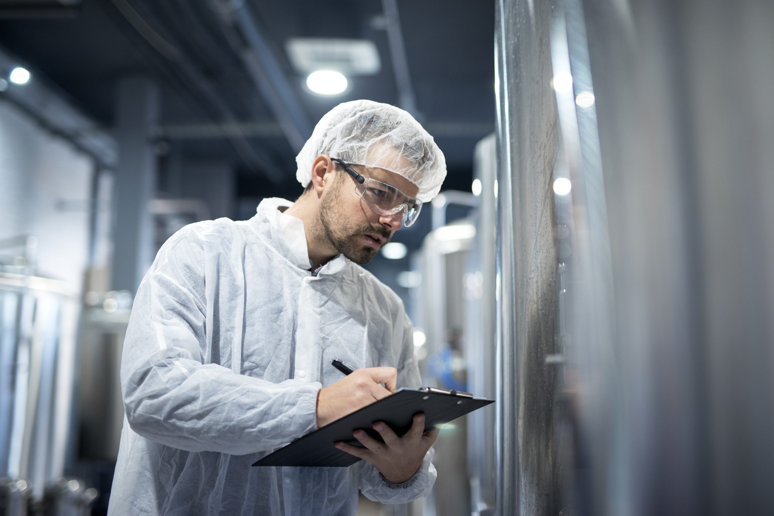 Especialista da Dourosafe em uniforme de proteção branco e óculos de segurança a realizar o registo técnico de monitorização num tanque industrial, representando o controlo de qualidade e segurança.