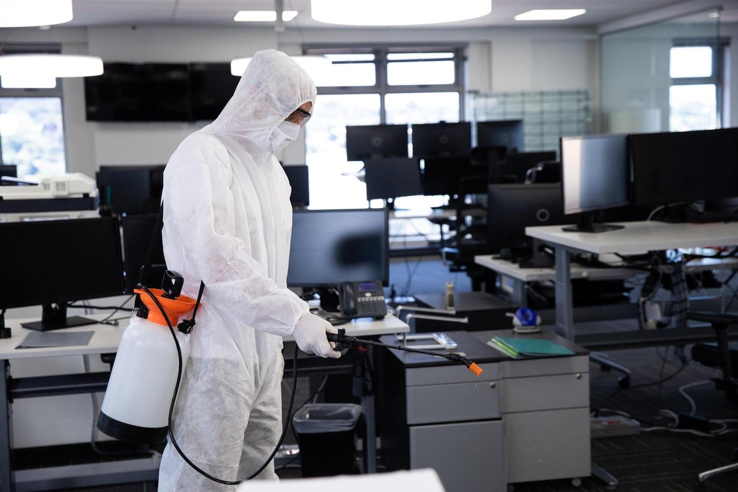 Técnico da Dourosafe em fato de proteção branco e máscara a realizar a pulverização para controlo de pragas num escritório moderno com secretárias e computadores desinfetados.