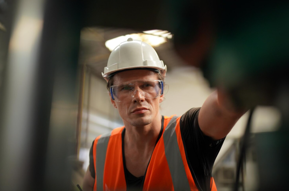 Especialista da Dourosafe focado, com capacete de proteção branco e colete de alta visibilidade laranja, a monitorizar e avaliar riscos de acidentes num ambiente de trabalho exigente.