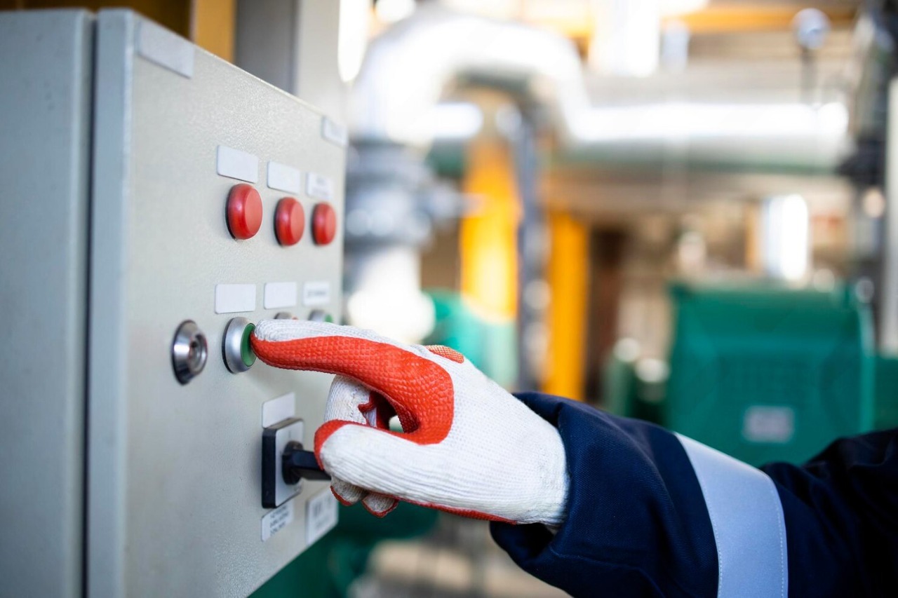 Close-up da mão de um operador industrial com luva de proteção laranja e branca a acionar o botão verde num painel de controlo elétrico, simbolizando a segurança operacional e HST.